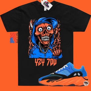 May include: Black t-shirt with a graphic of a skull wearing a blue hoodie and a gold chain. The text "424 700" is printed below the graphic. A pair of blue and orange sneakers are shown below the t-shirt.
