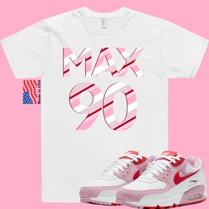 May include: White t-shirt with the words "MAX 90" in pink and red stripes.  Pink and white Nike Air Max 90 sneakers with red accents.