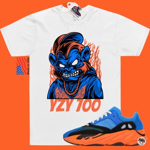 May include: White t-shirt with a blue and orange cartoon monkey graphic and the text "YZY 700". A pair of blue and orange Yeezy 700 sneakers are shown below the t-shirt.