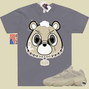 May include: A gray t-shirt with a cartoon bear wearing a chain and a pendant that says "YZY". The bear has a white fluffy head and a stitched-up face. The t-shirt is paired with a pair of beige sneakers with a mesh upper and a chunky sole.