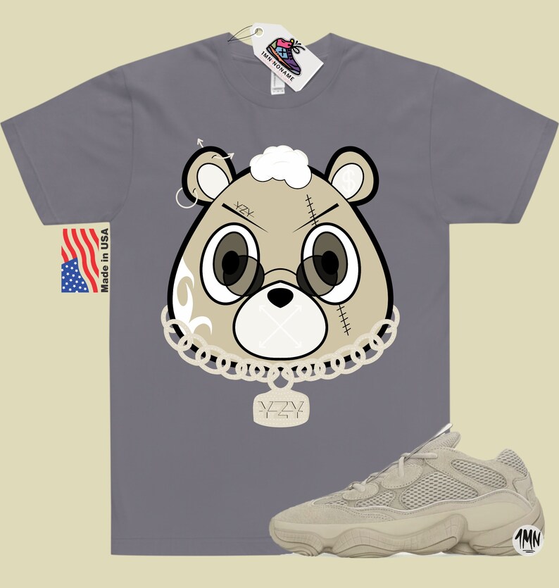 May include: A gray t-shirt with a cartoon bear wearing a chain and a pendant that says "YZY". The bear has a white fluffy head and a stitched-up face. The t-shirt is paired with a pair of beige sneakers with a mesh upper and a chunky sole.