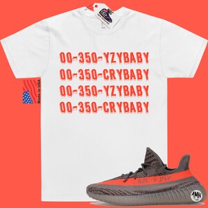 Peut inclure: T-shirt blanc avec du texte rouge qui dit "00-350-YZYBABY" et "00-350-CRYBABY" r&eacute;p&eacute;t&eacute; deux fois. Une paire de baskets grises et orange est repr&eacute;sent&eacute;e sous le t-shirt.