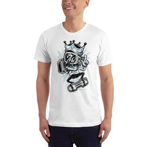 Puede incluir: Camiseta blanca con una ilustraci&oacute;n de dibujos animados de un personaje que lleva una corona y un casco, sosteniendo una patineta y apuntando con un dedo. El personaje est&aacute; en una pose de patinaje.