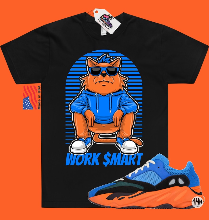 Op de afbeelding: Zwart T-shirt met een cartoon oranje kat die een zonnebril en een blauwe hoodie draagt. De kat zit met de tekst "WORK $MART" eronder. Een paar blauwe en oranje sneakers bevinden zich in de rechterbenedenhoek van de afbeelding.