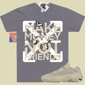 K&ouml;nnte beinhalten: Graues T-Shirt mit dem Text "MAKE MONEY NOT FRIENDS" in wei&szlig;en umrandeten Buchstaben. Ein Schlange, ein Schmetterling und ein Dollar-Schein sind in das Design integriert. Ein Paar beigefarbene Sneaker mit Mesh-Obermaterial sind unter dem T-Shirt zu sehen.