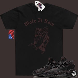 K&ouml;nnte beinhalten: Schwarzes T-Shirt mit dem Text "Make It Rain" in burgunderfarbener Schrift. Auf dem Shirt ist eine burgunderfarbene Illustration einer Hand, die Geld in einer betenden Position h&auml;lt. Unter dem Shirt befindet sich ein Paar schwarze und graue Sneaker.