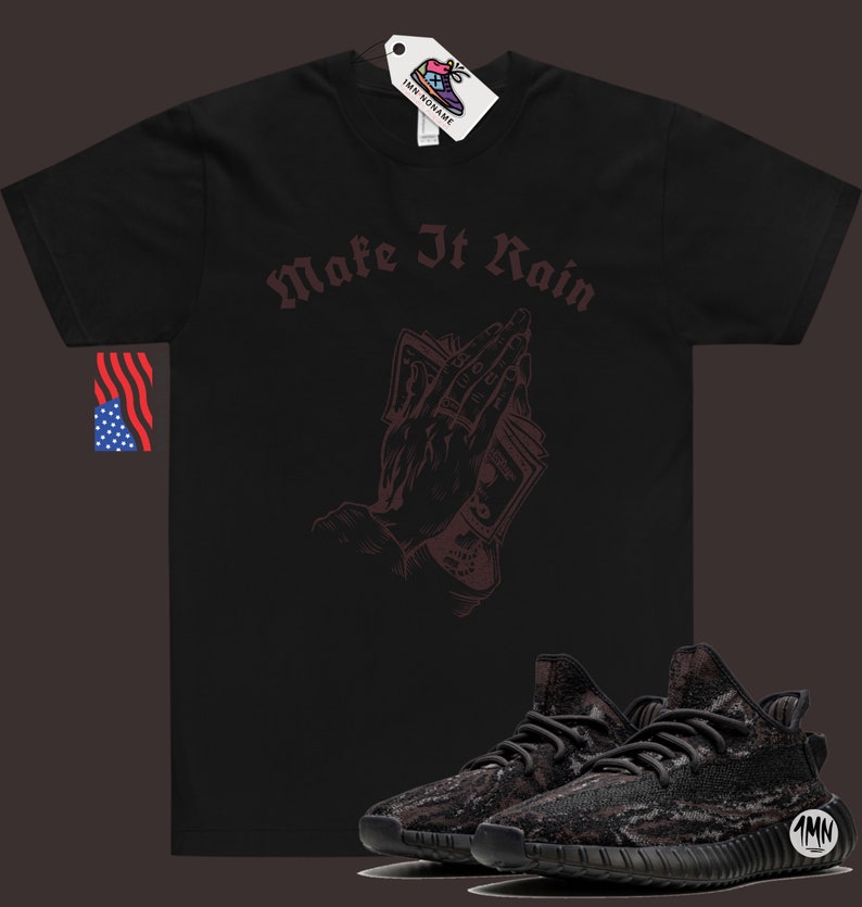 K&ouml;nnte beinhalten: Schwarzes T-Shirt mit dem Text "Make It Rain" in burgunderfarbener Schrift. Auf dem Shirt ist eine burgunderfarbene Illustration einer Hand, die Geld in einer betenden Position h&auml;lt. Unter dem Shirt befindet sich ein Paar schwarze und graue Sneaker.