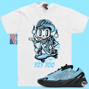 May include: A white t-shirt with a cartoon skeleton wearing a blue hat and jacket, skateboarding. The text "YZY 700" is printed below the image. A pair of blue and black Yeezy 700 sneakers are also pictured.