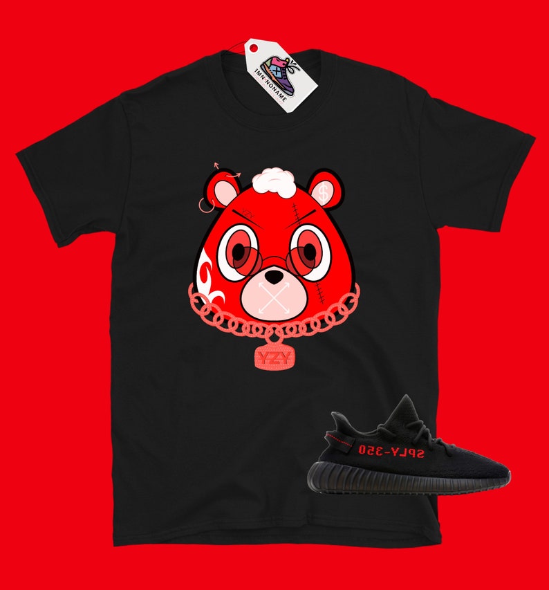 Puede incluir: Camiseta negra con un oso de dibujos animados rojo que lleva una cadena con un colgante YZY. El oso tiene ojos rojos y la boca cosida. Debajo de la camiseta se muestra un par de zapatillas Yeezy negras y rojas.
