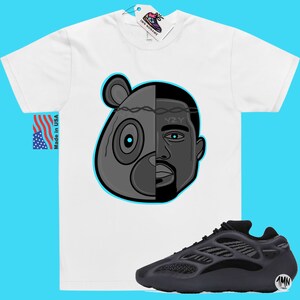 Op de afbeelding: Een wit T-shirt met een zwart-witte illustratie van een berenkop met een menselijk gezicht aan de rechterkant. De tekst "YZY" staat op het voorhoofd. Het T-shirt wordt gecombineerd met zwarte en grijze Yeezy 700 sneakers.