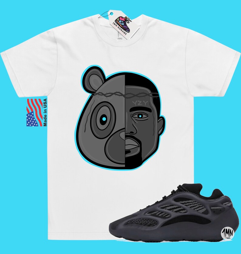 Op de afbeelding: Een wit T-shirt met een zwart-witte illustratie van een berenkop met een menselijk gezicht aan de rechterkant. De tekst "YZY" staat op het voorhoofd. Het T-shirt wordt gecombineerd met zwarte en grijze Yeezy 700 sneakers.