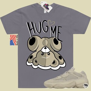 Puede incluir: Una camiseta gris con un oso de peluche de dibujos animados que lleva un coraz&oacute;n y el texto "Hug Me". Tambi&eacute;n se ve un par de zapatillas beige con una parte superior de malla y una suela gruesa.
