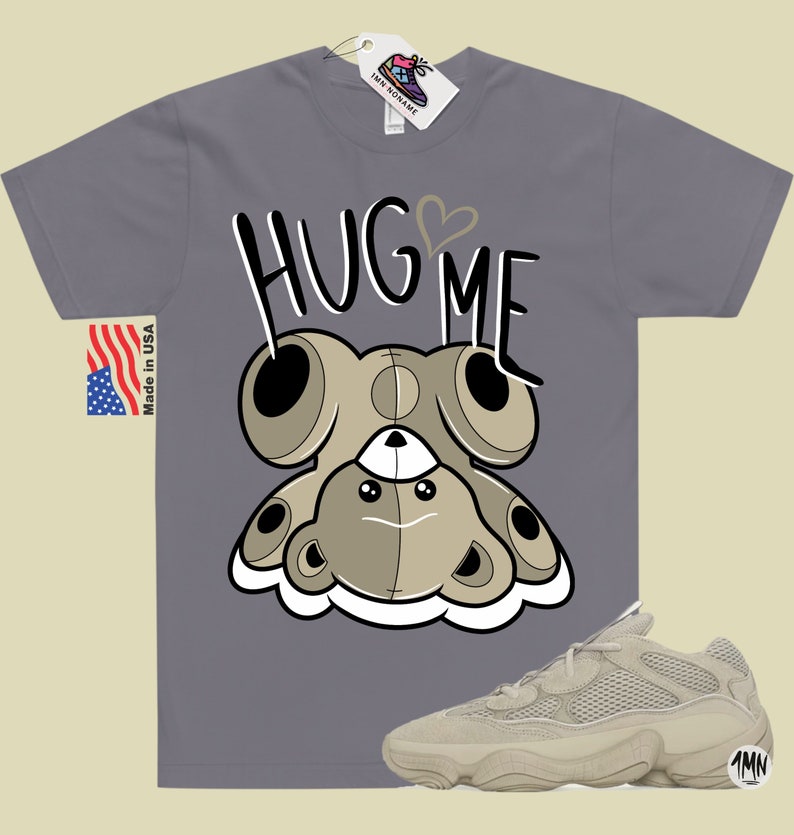 Puede incluir: Una camiseta gris con un oso de peluche de dibujos animados que lleva un coraz&oacute;n y el texto "Hug Me". Tambi&eacute;n se ve un par de zapatillas beige con una parte superior de malla y una suela gruesa.