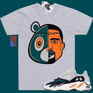 May include: A grey t-shirt with a graphic of a cartoon bear and Kanye West's face. The graphic is split down the middle, with the bear on the left and Kanye on the right. The bear is wearing a crown of thorns. The t-shirt is paired with a pair of white, black, and orange sneakers with the text "YZY" on the side.
