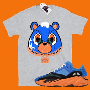 May include: A grey t-shirt with a blue bear graphic wearing a gold chain with a YZY pendant. The bear has a dollar sign on its head and a stitched-up mouth with an X. The t-shirt is paired with blue and orange YZY sneakers.