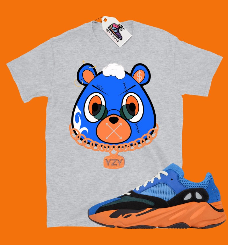May include: A grey t-shirt with a blue bear graphic wearing a gold chain with a YZY pendant. The bear has a dollar sign on its head and a stitched-up mouth with an X. The t-shirt is paired with blue and orange YZY sneakers.