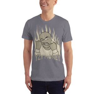 May include: A gray t-shirt with a graphic of a person wearing a ski mask and making a hand gesture. The graphic is in a beige color and has flames behind it. The text "YZY MONEY" is printed below the graphic.