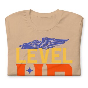 K&ouml;nnte beinhalten: Ein beiges T-Shirt mit einer blauen Adlergrafik und dem Text "LEVEL UP" in Gelb und Orange.