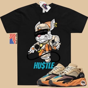 May include: Black t-shirt with a cartoon rabbit wearing sunglasses and a hat, holding a microphone and a spray can. The text "HUSTLE" is printed below the rabbit. A pair of tan, orange, and blue sneakers are shown below the t-shirt.