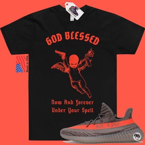 Puede incluir: Camiseta negra con texto rojo que dice "GOD BLESSED" y un gr&aacute;fico de un querub&iacute;n sosteniendo una pistola y un c&oacute;ctel molotov. El texto "Now And forever Under Your Spell" est&aacute; debajo del gr&aacute;fico. Un par de zapatillas Yeezy grises y naranjas se encuentran en la esquina inferior derecha de la imagen.