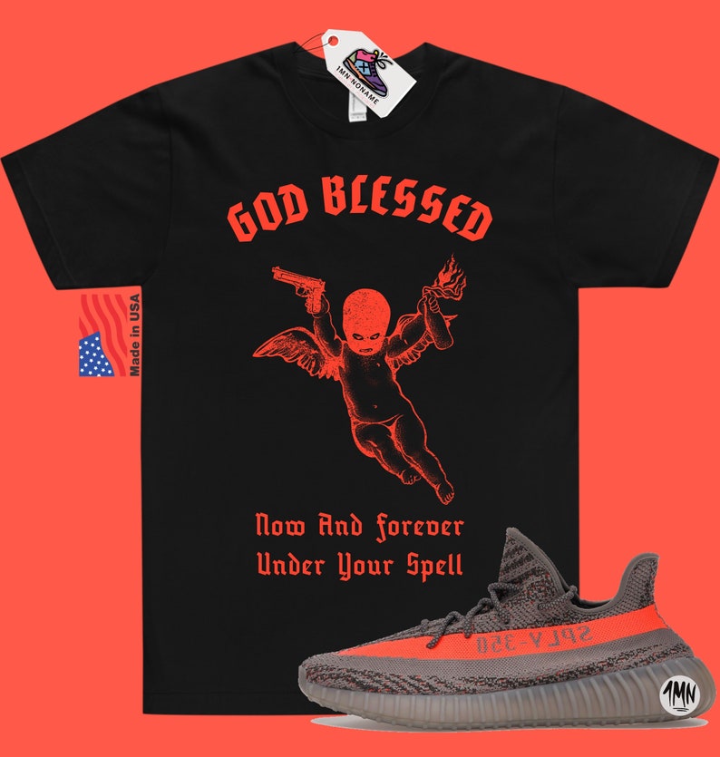 Puede incluir: Camiseta negra con texto rojo que dice "GOD BLESSED" y un gr&aacute;fico de un querub&iacute;n sosteniendo una pistola y un c&oacute;ctel molotov. El texto "Now And forever Under Your Spell" est&aacute; debajo del gr&aacute;fico. Un par de zapatillas Yeezy grises y naranjas se encuentran en la esquina inferior derecha de la imagen.