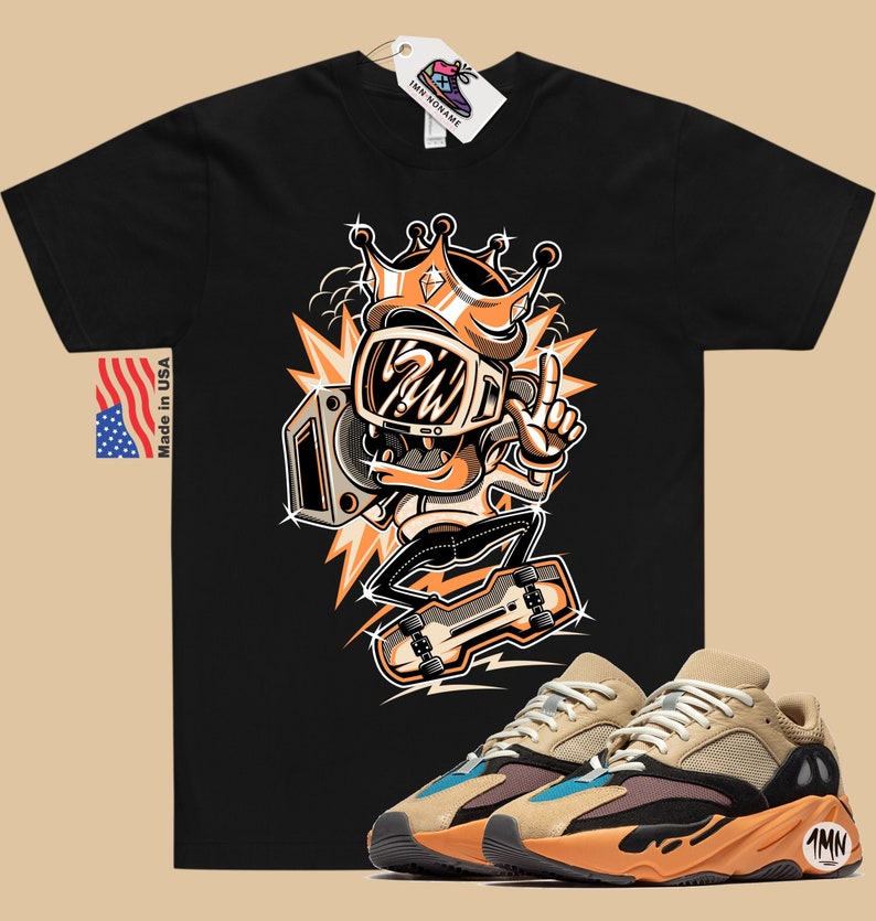 Może przedstawiać: Czarny T-shirt z kresk&oacute;wkową ilustracją postaci w koronie trzymającej boombox. Postać jeździ na deskorolce. Pod T-shirtem widoczna jest para beżowych, pomarańczowych i niebieskich but&oacute;w sportowych.