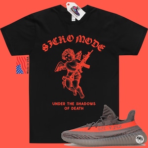 May include: Black t-shirt with a red graphic of a cherub holding a gun. The text "SICKO MODE" is above the cherub and "UNDER THE SHADOWS OF DEATH" is below. A pair of gray and orange Yeezy sneakers are in the bottom right corner of the image.