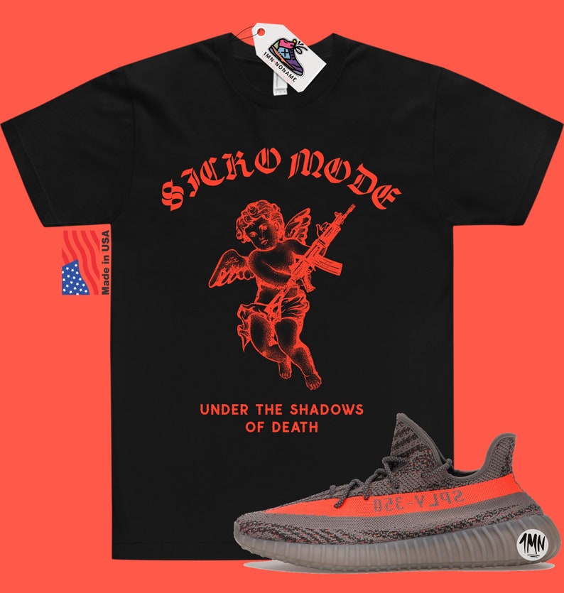 May include: Black t-shirt with a red graphic of a cherub holding a gun. The text "SICKO MODE" is above the cherub and "UNDER THE SHADOWS OF DEATH" is below. A pair of gray and orange Yeezy sneakers are in the bottom right corner of the image.