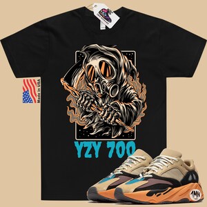 May include: Black t-shirt with a graphic of a hooded figure wearing a gas mask and holding a scythe. The text "YZY 700" is printed below the graphic. A pair of tan and orange sneakers with blue accents are shown in the bottom right corner of the image.