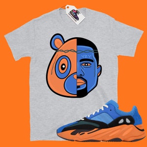 Pode incluir: Uma camiseta cinza com um gr&aacute;fico de uma cabe&ccedil;a de urso e o rosto de Kanye West. O gr&aacute;fico &eacute; dividido ao meio por uma coroa de arame farpado. A camiseta &eacute; combinada com t&ecirc;nis laranja e azul com o texto "YZY" no lado.