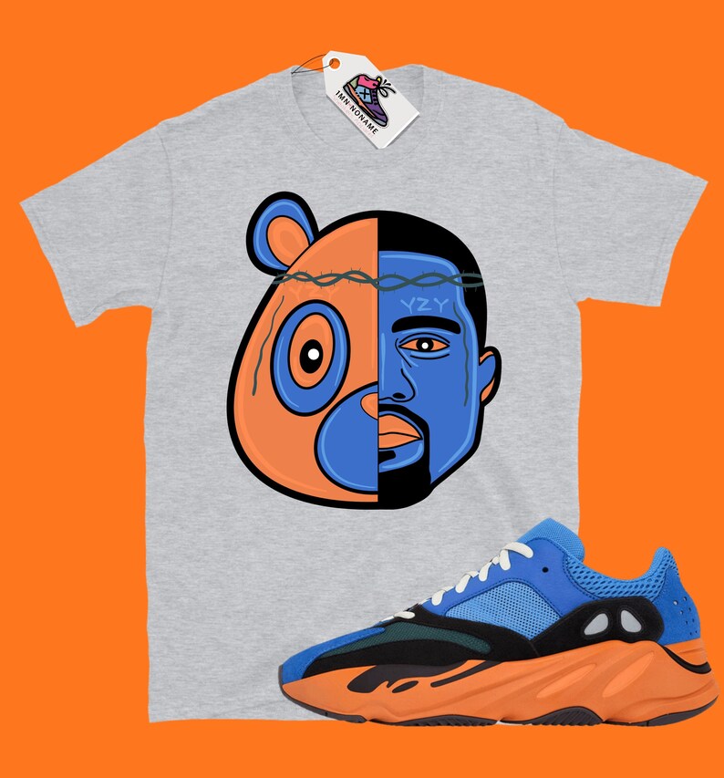 Pode incluir: Uma camiseta cinza com um gr&aacute;fico de uma cabe&ccedil;a de urso e o rosto de Kanye West. O gr&aacute;fico &eacute; dividido ao meio por uma coroa de arame farpado. A camiseta &eacute; combinada com t&ecirc;nis laranja e azul com o texto "YZY" no lado.