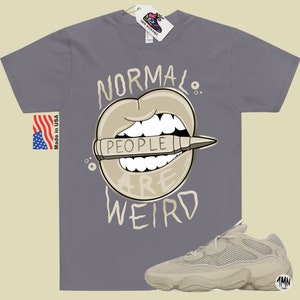 Pode incluir: Uma camiseta cinza com o texto "Normal People Are Weird" em preto e branco. O texto &eacute; cercado por uma imagem de desenho animado de uma boca com um l&aacute;pis dentro. O l&aacute;pis tem a palavra "People" escrita nele. Tamb&eacute;m &eacute; mostrado um par de t&ecirc;nis bege.