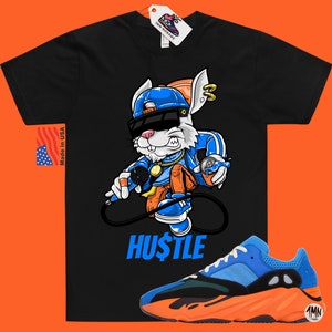 Puede incluir: Camiseta negra con un conejo de dibujos animados que lleva un sombrero azul, gafas de sol y una cadena de oro, sosteniendo un micr&oacute;fono y una lata de spray. El texto "HUSTLE" est&aacute; debajo del conejo. Un par de zapatillas azules y naranjas se encuentran en la esquina inferior derecha de la imagen.
