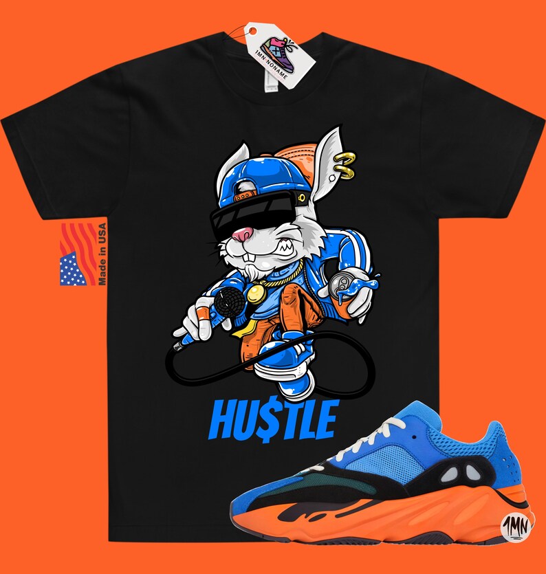 Puede incluir: Camiseta negra con un conejo de dibujos animados que lleva un sombrero azul, gafas de sol y una cadena de oro, sosteniendo un micr&oacute;fono y una lata de spray. El texto "HUSTLE" est&aacute; debajo del conejo. Un par de zapatillas azules y naranjas se encuentran en la esquina inferior derecha de la imagen.