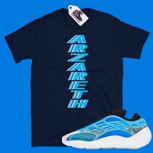 Pu&ograve; includere: Una t-shirt blu navy con la parola "ARZARETH" stampata verticalmente in strisce blu e bianche. Un paio di scarpe da ginnastica blu e bianche con suola bianca.