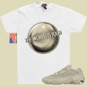 May include: White t-shirt with a gold sphere graphic and the text "The World Is Yours". A pair of beige sneakers with a mesh upper and a thick sole are also pictured.