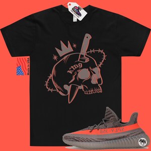 May include: Black t-shirt with a graphic of a skull with a knife through it and a crown on top. The text "King" is on the skull. The t-shirt is paired with a pair of grey and orange sneakers with the text "Yeezy 350" on the side.