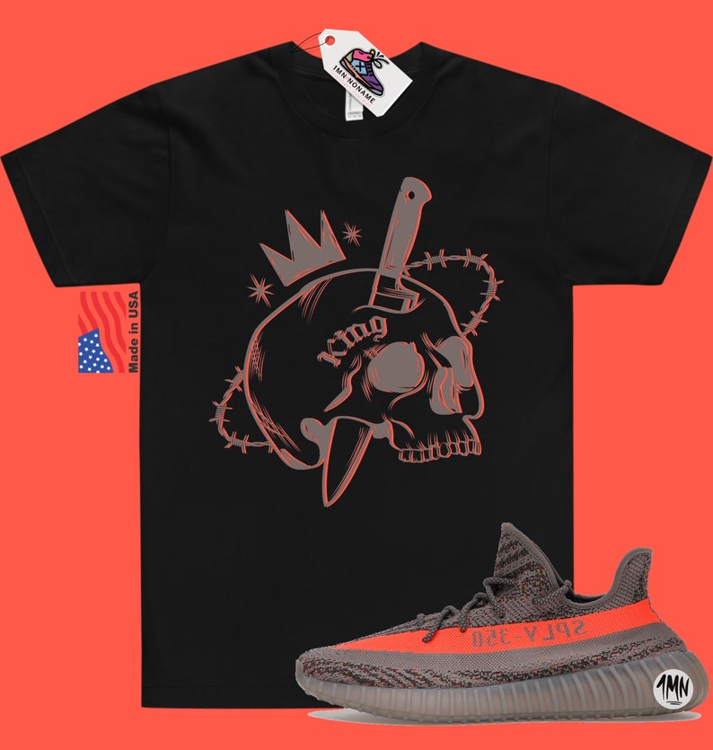May include: Black t-shirt with a graphic of a skull with a knife through it and a crown on top. The text "King" is on the skull. The t-shirt is paired with a pair of grey and orange sneakers with the text "Yeezy 350" on the side.