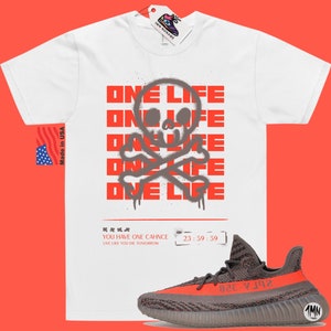 Puede incluir: Camiseta blanca con un gr&aacute;fico rojo de calavera y huesos cruzados y el texto "ONE LIFE" repetido varias veces. El texto est&aacute; en estilo de pintura en aerosol. Debajo del gr&aacute;fico est&aacute; el texto "YOU HAVE ONE CHANCE LIVE LIKE YOU DIE TOMORROW" y los n&uacute;meros "23:59:59". Un par de zapatillas grises y rojas se muestra debajo de la camiseta.