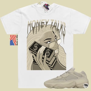 May include: White t-shirt with a graphic of a woman with blonde hair wearing glasses and holding a stack of money to her ear. The text "MONEY TALKS" is above the woman. A pair of beige sneakers are in the bottom right corner of the image.