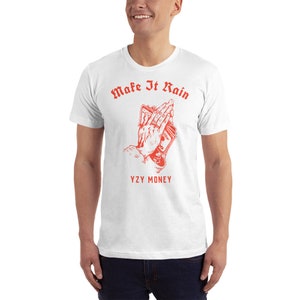 Puede incluir: Camiseta blanca con un gr&aacute;fico rojo de manos sosteniendo dinero en posici&oacute;n de oraci&oacute;n. El texto "Make It Rain" est&aacute; por encima del gr&aacute;fico y "YZY MONEY" est&aacute; por debajo.