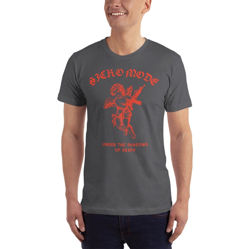 May include: A dark gray t-shirt with a red graphic of a cherub holding a rifle. The text "SICKO MODE" is above the cherub and "UNDER THE SHADOWS OF DEATH" is below the cherub.