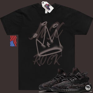 Puede incluir: Camiseta negra con una corona pintada con aerosol y la palabra "ROCK" en blanco. Un par de zapatillas negras y marrones con estampado de camuflaje se muestran debajo de la camiseta.