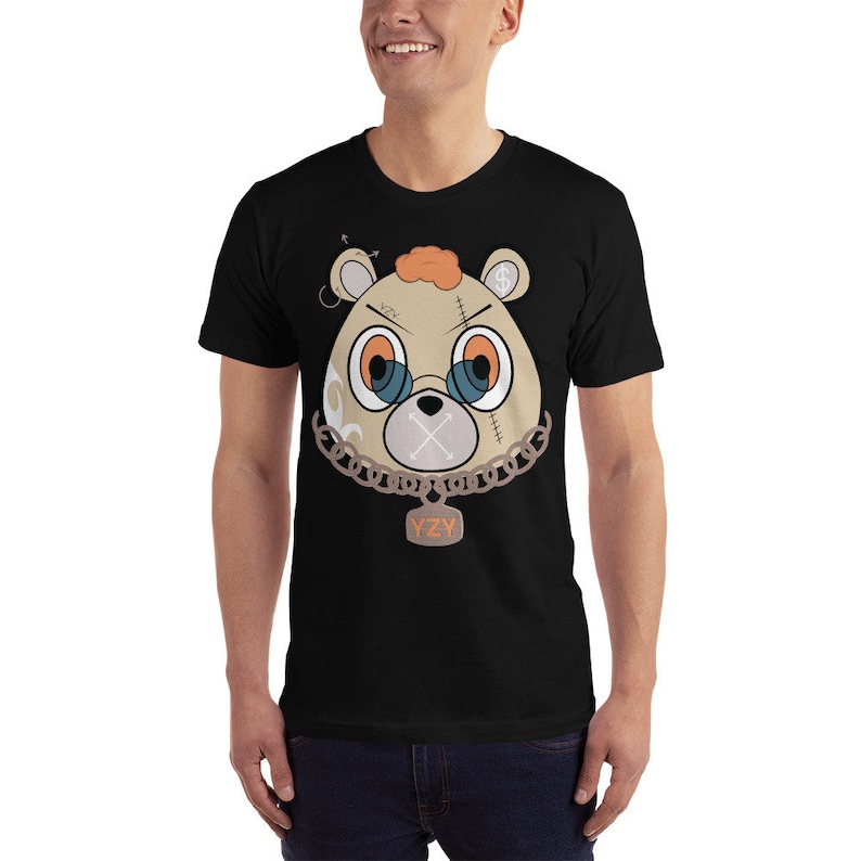 May include: A black t-shirt featuring a cartoon image of a brown bear with a gold chain and a tag that reads "YZY". The bear has a stitched-up face with a blue eye patch and a black X over its mouth.