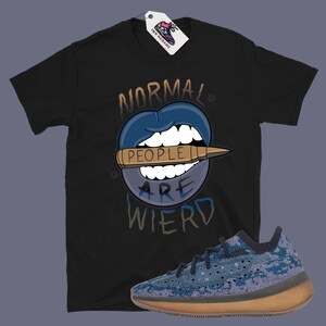 Op de afbeelding: Zwart T-shirt met een afbeelding van een blauwe mond met een kogel waarop "people" staat en de tekst "Normal People Are Wierd". Er is ook een paar blauwe en bruine sneakers te zien.
