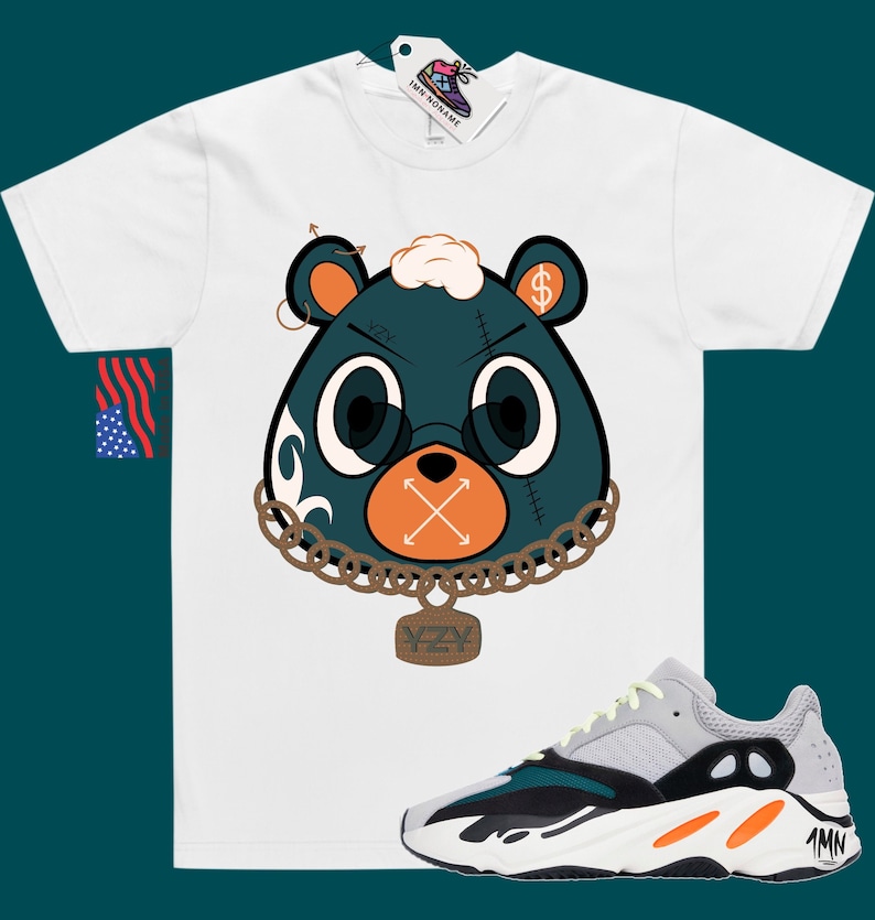 May include: White t-shirt featuring a cartoon illustration of a green bear wearing a gold chain with a YZY pendant. The bear has a dollar sign on its ear and an X over its mouth. The t-shirt is paired with grey, green, and orange trainers.
