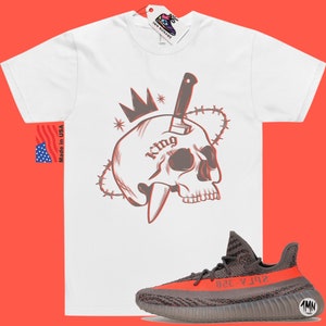 May include: White t-shirt with a graphic of a skull with a knife through it and a crown on top. The skull has the word "King" tattooed on it. The graphic is surrounded by barbed wire. The t-shirt is paired with a pair of grey and orange sneakers.