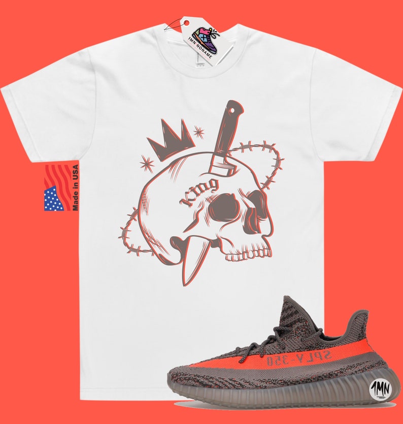 May include: White t-shirt with a graphic of a skull with a knife through it and a crown on top. The skull has the word "King" tattooed on it. The graphic is surrounded by barbed wire. The t-shirt is paired with a pair of grey and orange sneakers.