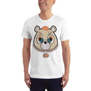 May include: White t-shirt featuring a cartoon image of a brown bear with a chain and a tag that reads "YZY". The bear has orange hair, blue eyes, and a stitched-up mouth.