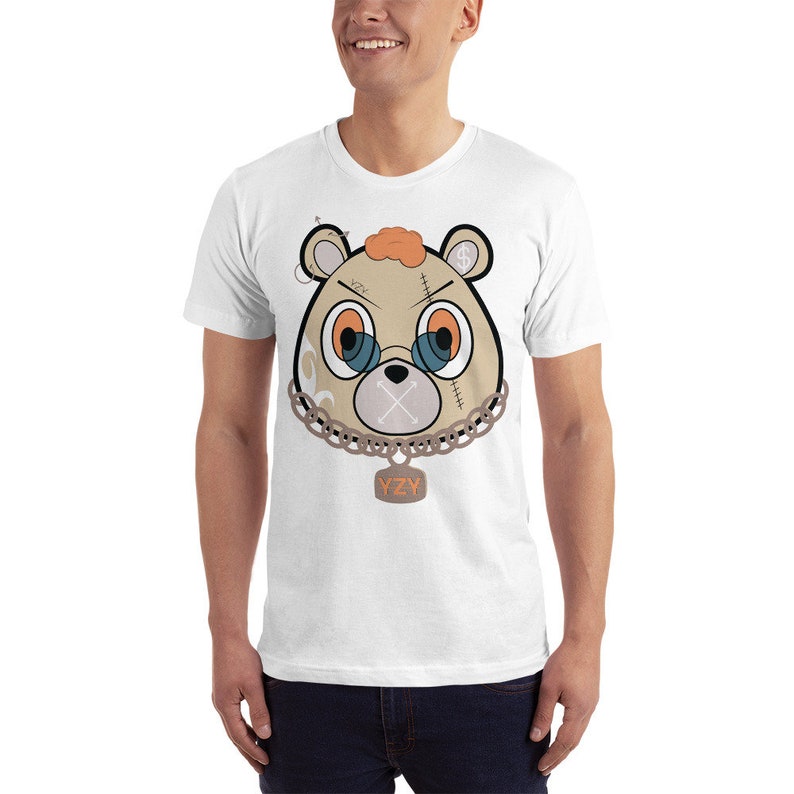 May include: White t-shirt featuring a cartoon image of a brown bear with a chain and a tag that reads "YZY". The bear has orange hair, blue eyes, and a stitched-up mouth.